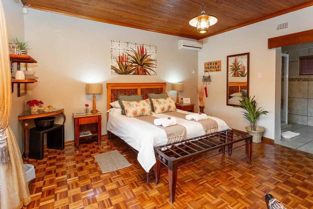 A well-lit bedroom with a wooden floor, a bed with multiple pillows, a bedside table with a lamp, and a large mirror on the wall.