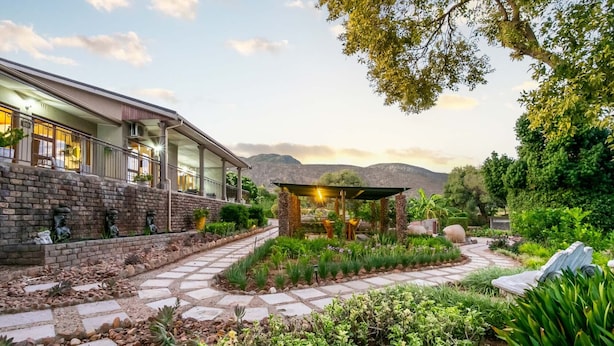 A modern building with a stone facade, a covered outdoor seating area, and a landscaped garden with a stone pathway. - Die Fonteine Guesthouse and Glamping (Oudtshoorn)