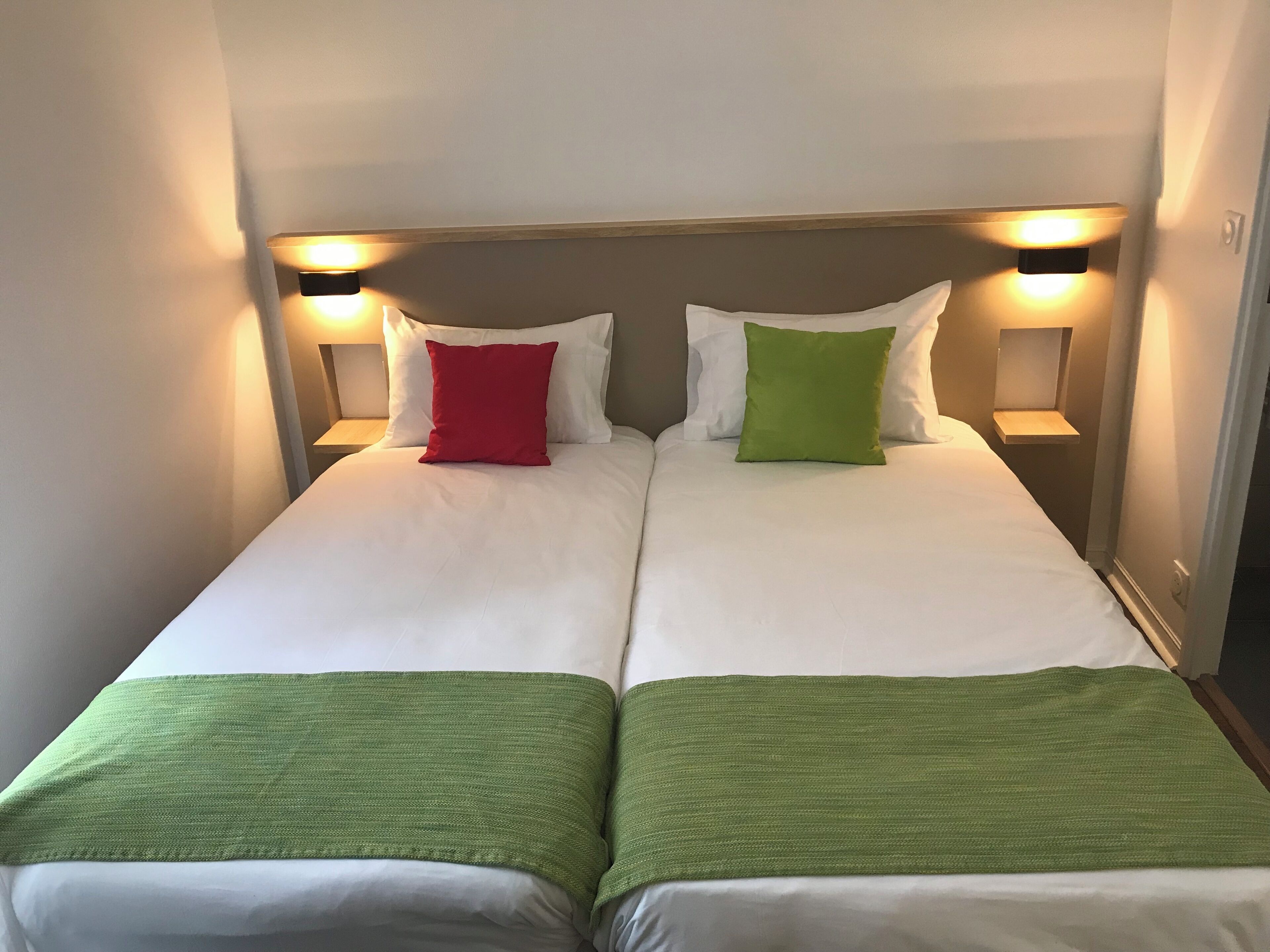 Ein Doppelbett mit weißer Bettwäsche, zwei rote und zwei grüne Kissen sowie zwei Nachttischlampen.
