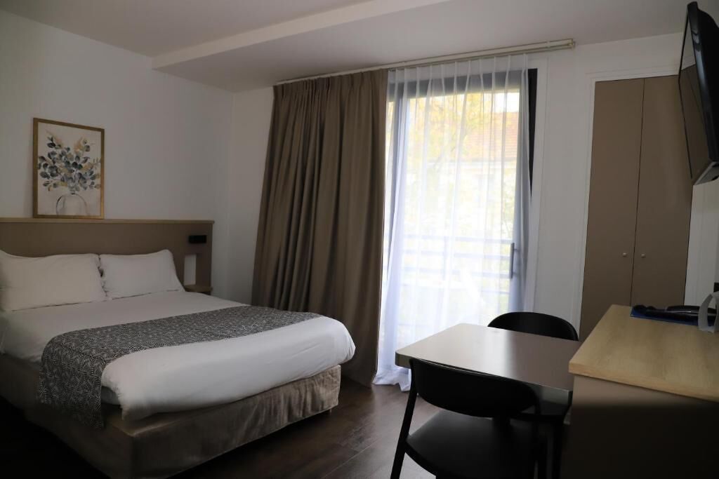 Habitación de hotel con cama, escritorio, silla, televisión y ventana con cortinas.