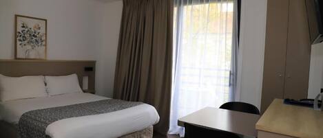 Habitación de hotel con cama, escritorio, silla, televisión y ventana con cortinas.