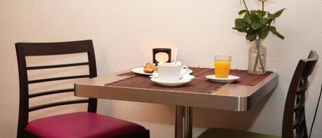 A dining table with a white cup, a glass of orange juice, and a small plate with a pastry. There are two chairs, one with a red cushion and one with a green cushion. A vase with two roses is on the table.