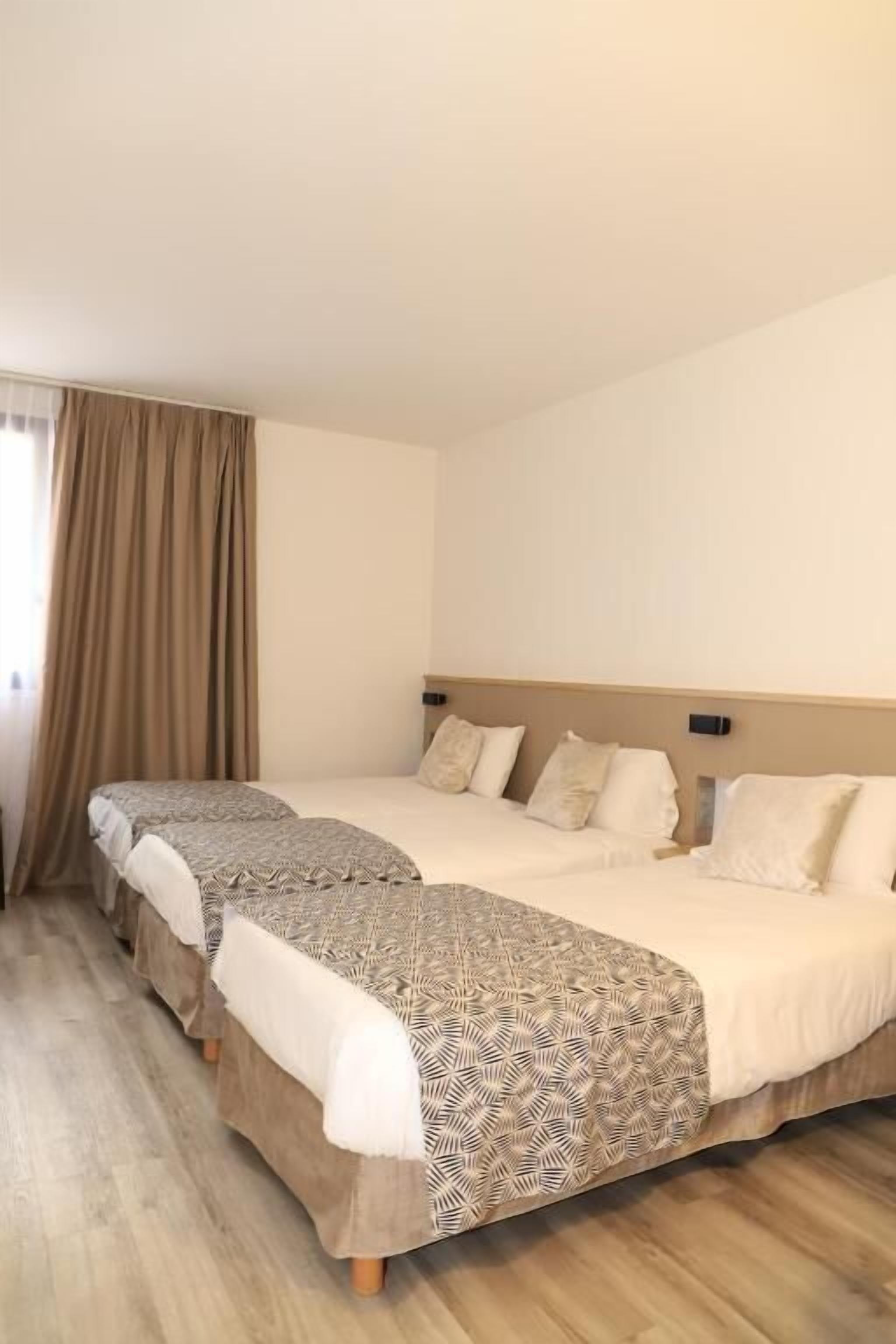 Habitación de hotel con dos camas, un ventanal grande con cortinas y suelo de madera.