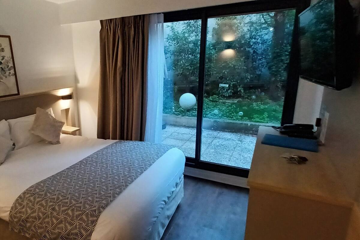 A hotel room with a bed, bedside lamps, a TV, and a view of a garden through a glass door.