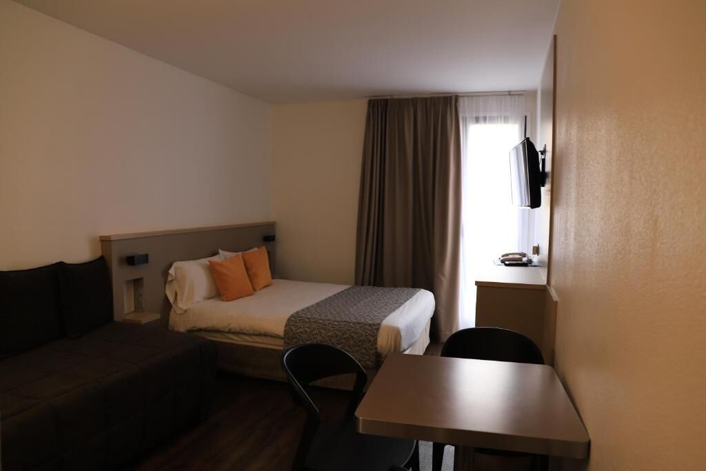 Habitación de hotel con cama, escritorio, silla, televisión y ventana con cortinas.