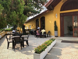 An outdoor cafe area with tables and chairs, a yellow building with a large door, and a tree providing shade.
