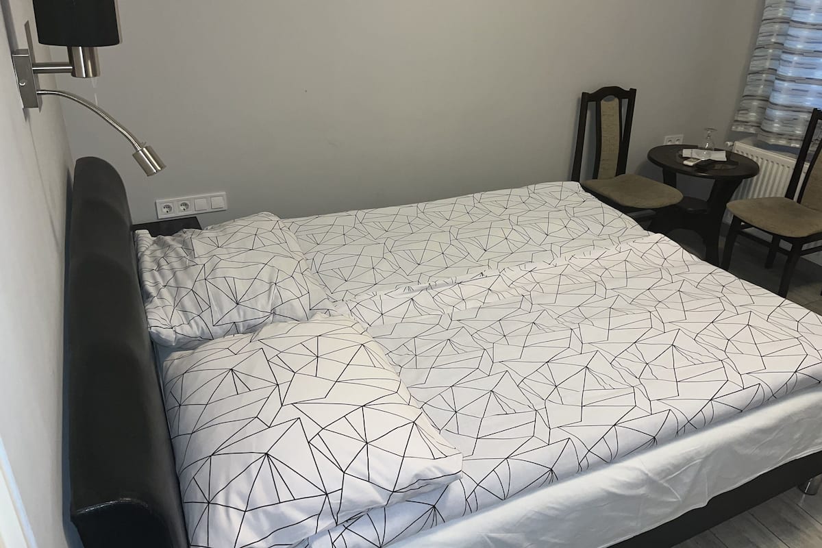 Un lit bien fait, recouvert d’une couvre-lit à motifs géométriques, une table de chevet avec une lampe et une chaise dans un petit espace adjacent.