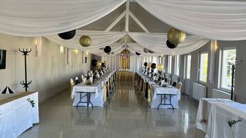 Un salón de banquetes con largas mesas dispuestas para un evento formal, cubiertas con tela blanca y detalles dorados, y amplias ventanas a lo largo de las paredes.