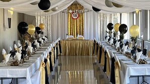 A long banquet hall with tables set for a formal event, draped with white and gold decorations, and a large number '18' sign in the background.
