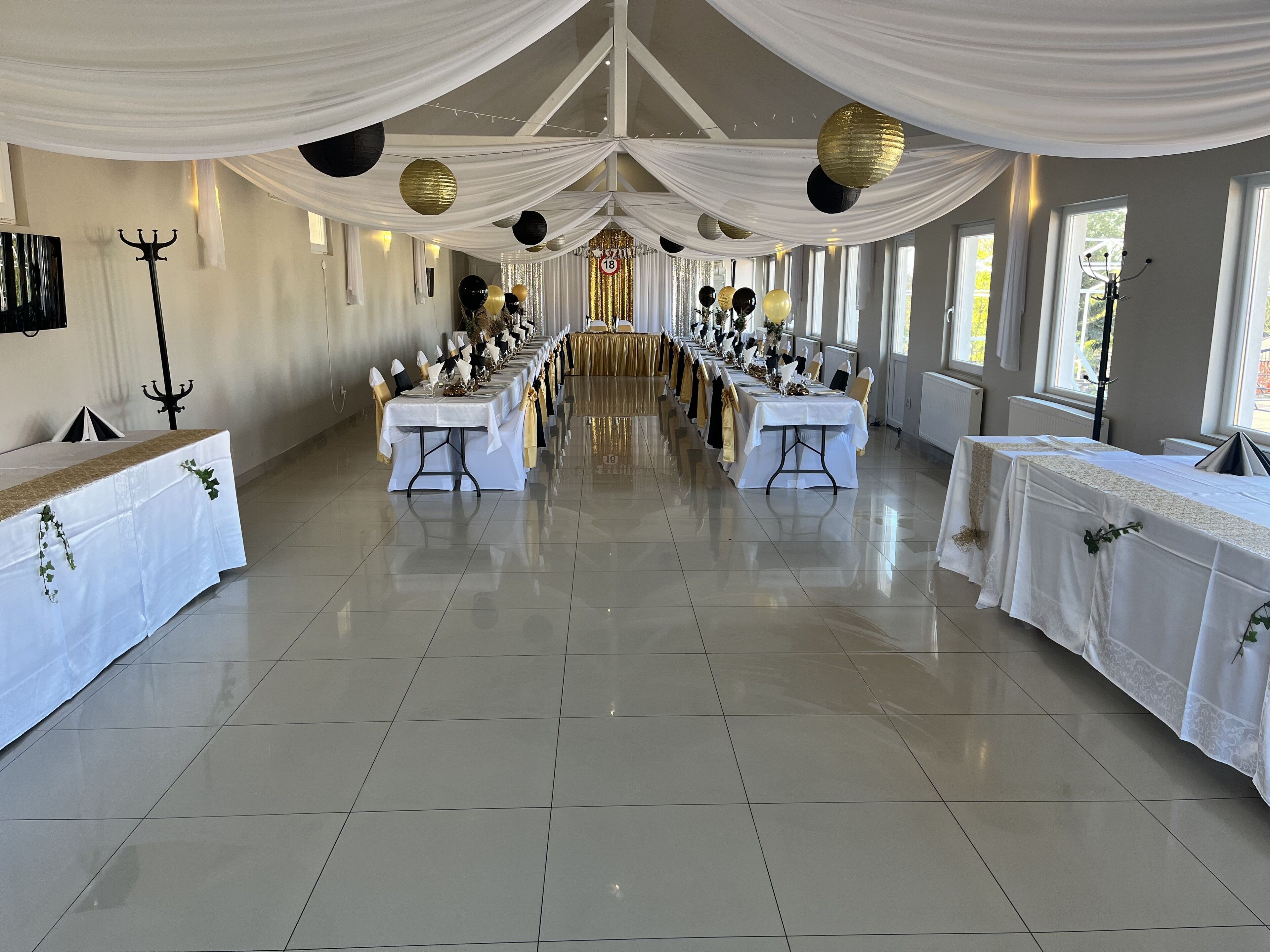 A banquet hall with long tables set for a formal event, draped with white fabric and gold accents, and large windows providing natural light.