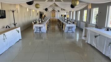 A banquet hall with long tables set for a formal event, draped with white fabric and gold accents, and large windows providing natural light.