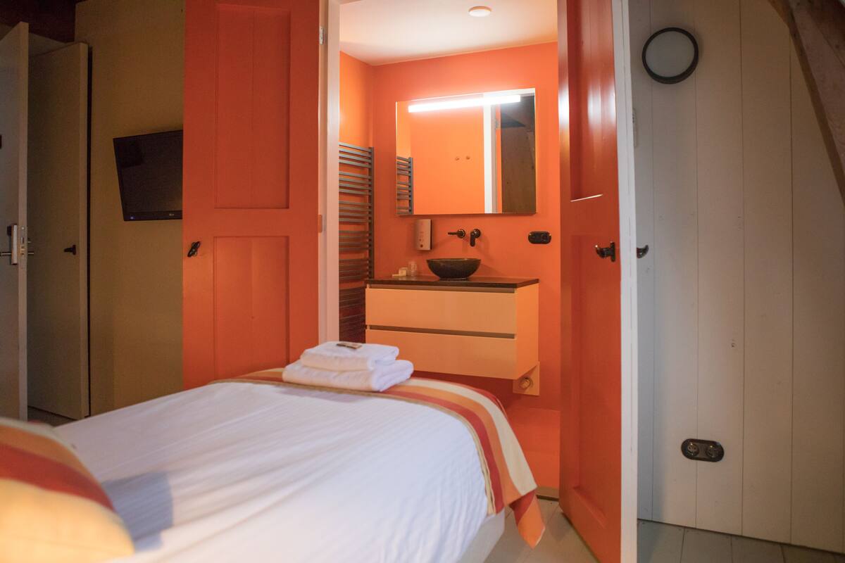 Una habitación de hotel con cama, baño con lavamanos y espejo, y una puerta.
