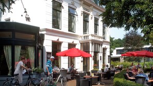 Un espace café en plein air, avec des meubles en osier et des parasols rouges, un vélo garé à proximité, et un bâtiment sur lequel on peut lire l’inscription « LUNIA ».