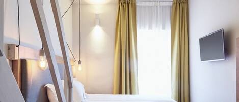 Una habitación de hotel con cama, televisor montado en la pared, mesita de noche con lámpara y cortinas en la ventana.