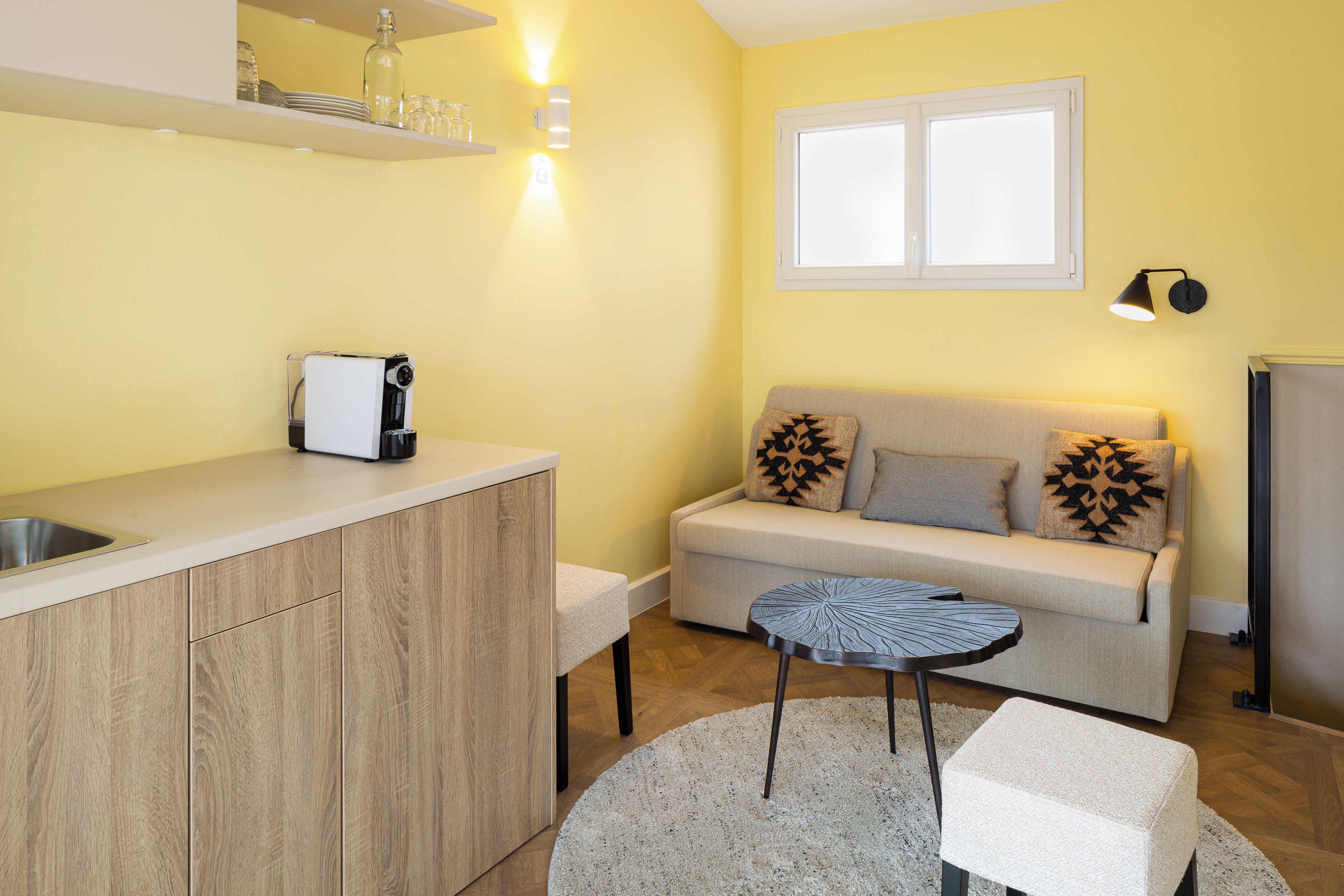 A compact kitchen with a built-in oven, a sink, and a countertop. There is a small round table with a chair, a sofa with patterned cushions, and a window above the sofa.