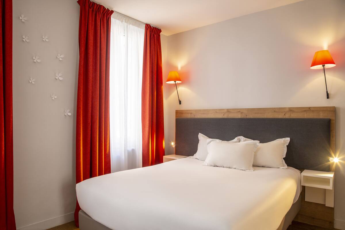 Une chambre d’hôtel avec un grand lit, deux lampes de chevet, une tête de lit en bois et une fenêtre avec des rideaux rouges.