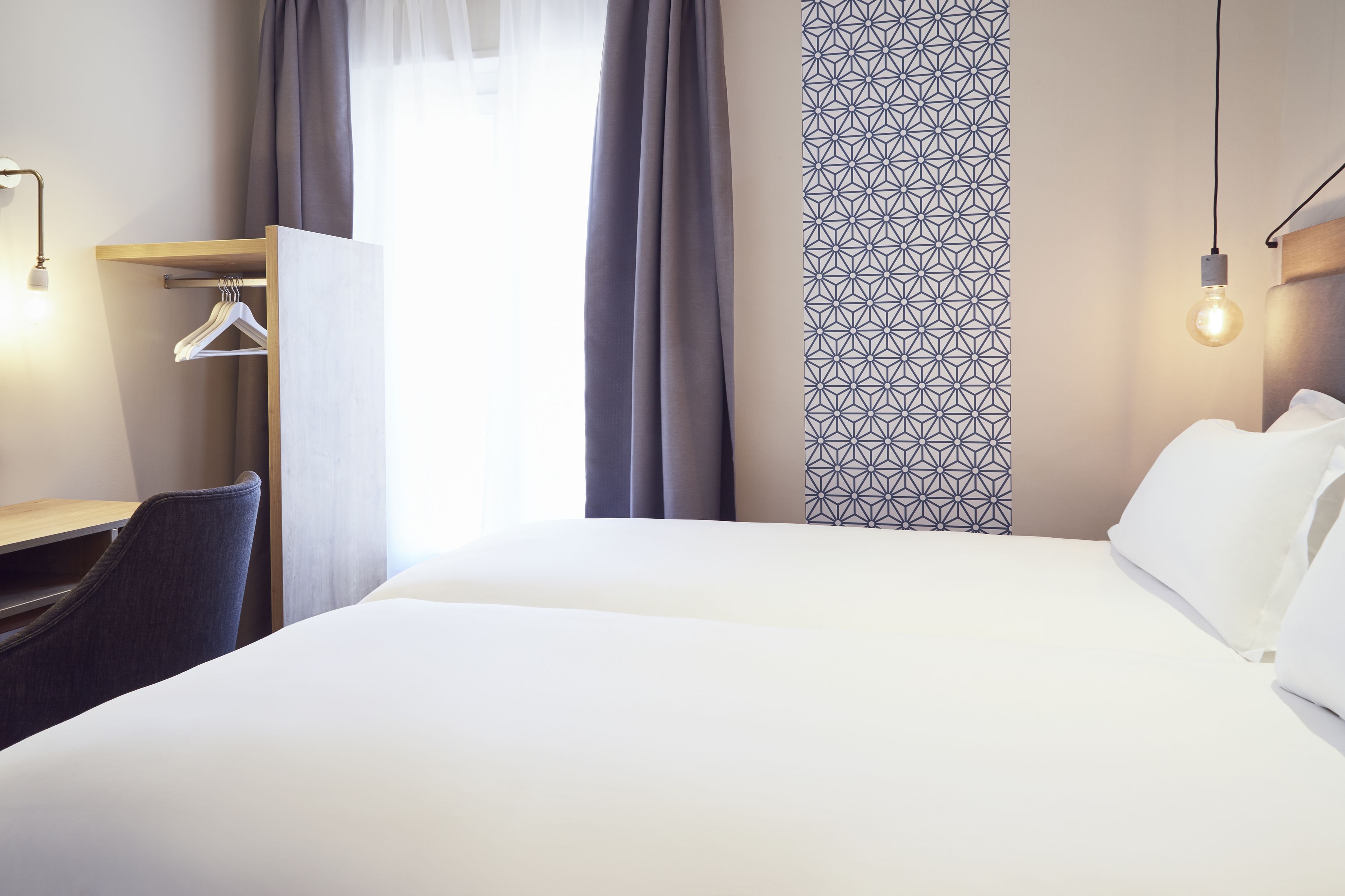 Une chambre d’hôtel avec un grand lit, un bureau avec une chaise, un porte-manteau et un rideau à motifs.