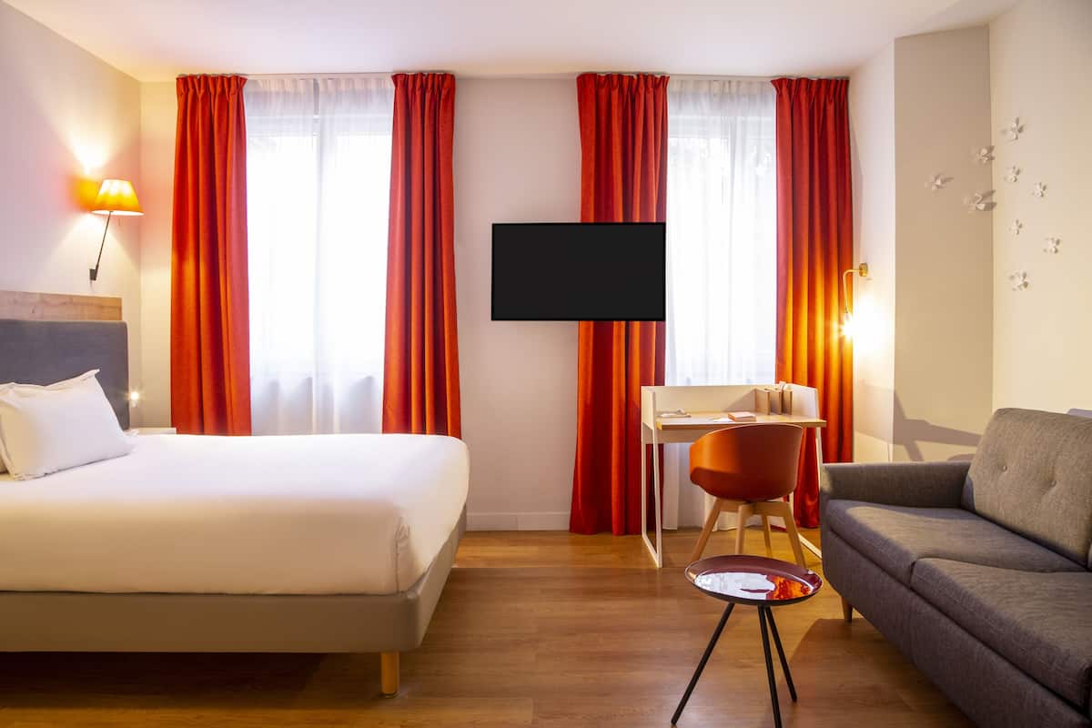 Une chambre d’hôtel avec un lit, un canapé, un bureau avec une chaise, un téléviseur fixé au mur et des rideaux rouges.
