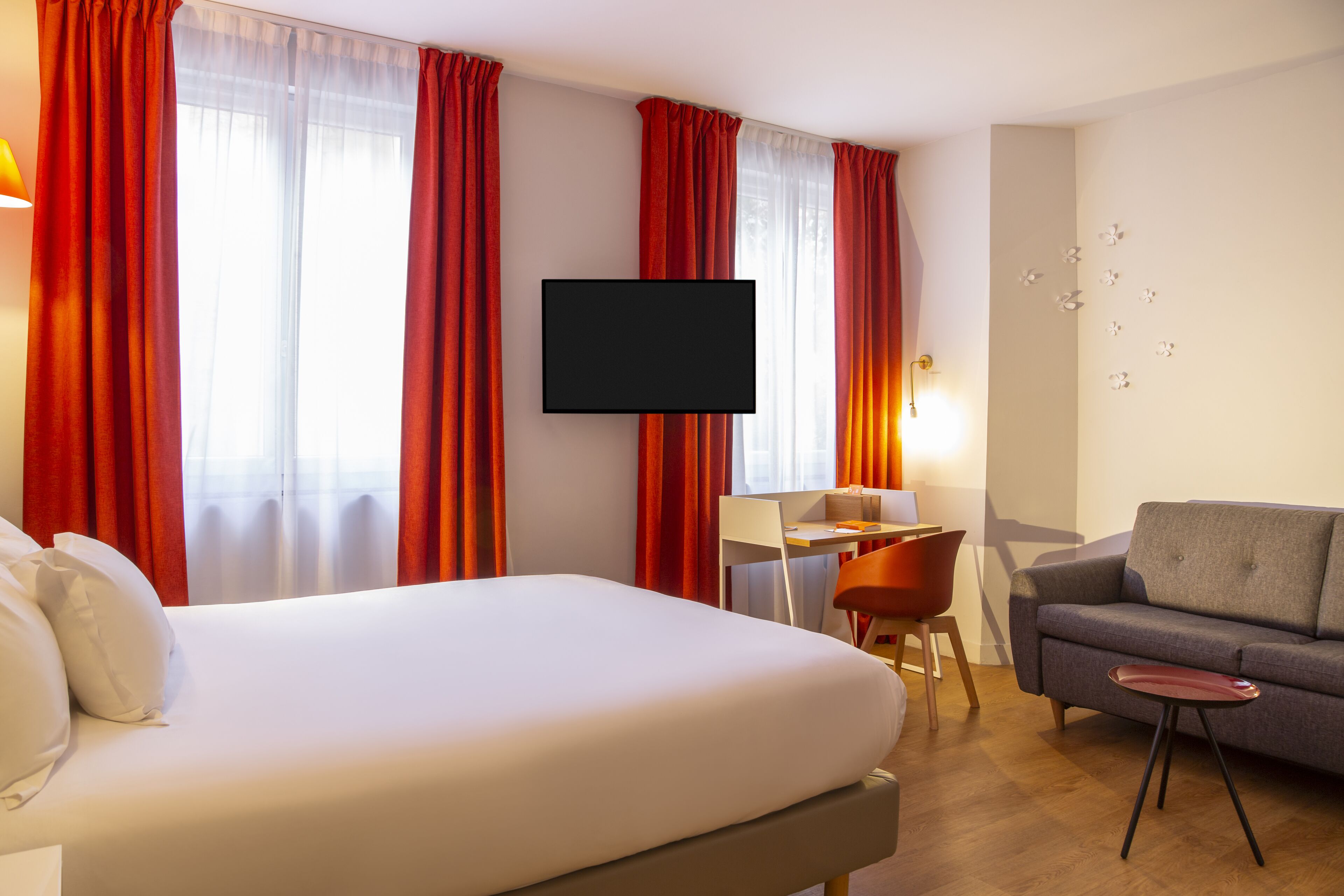 A hotel room with a bed, a desk, a chair, a sofa, and a TV mounted on the wall.