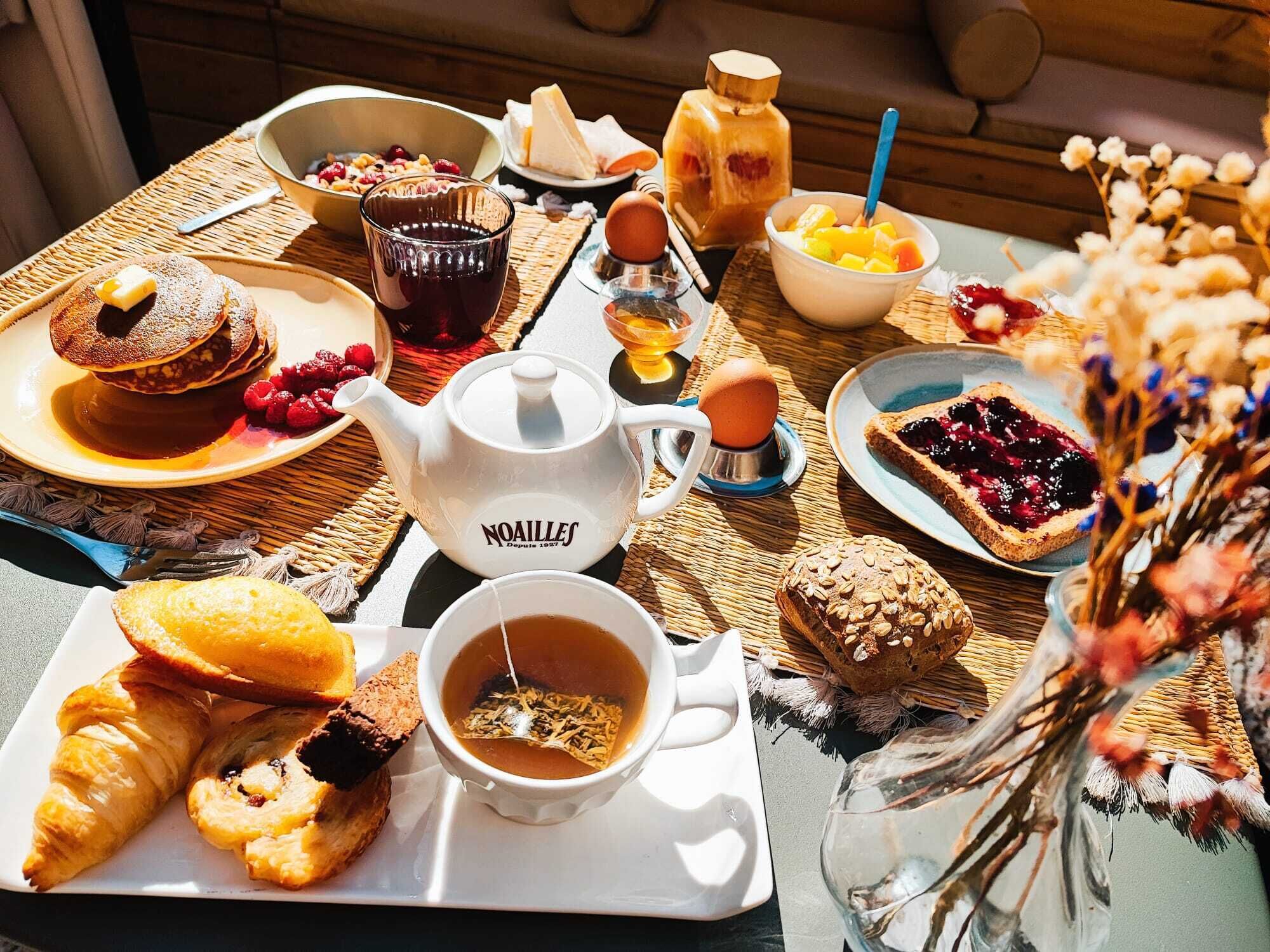 A breakfast table with pancakes, toast, croissants, a teapot, and a cup of tea.