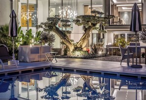Un área interior de piscina moderna con una gran palmera como centro, sillas de descanso y un comedor con mesas y sillas.
