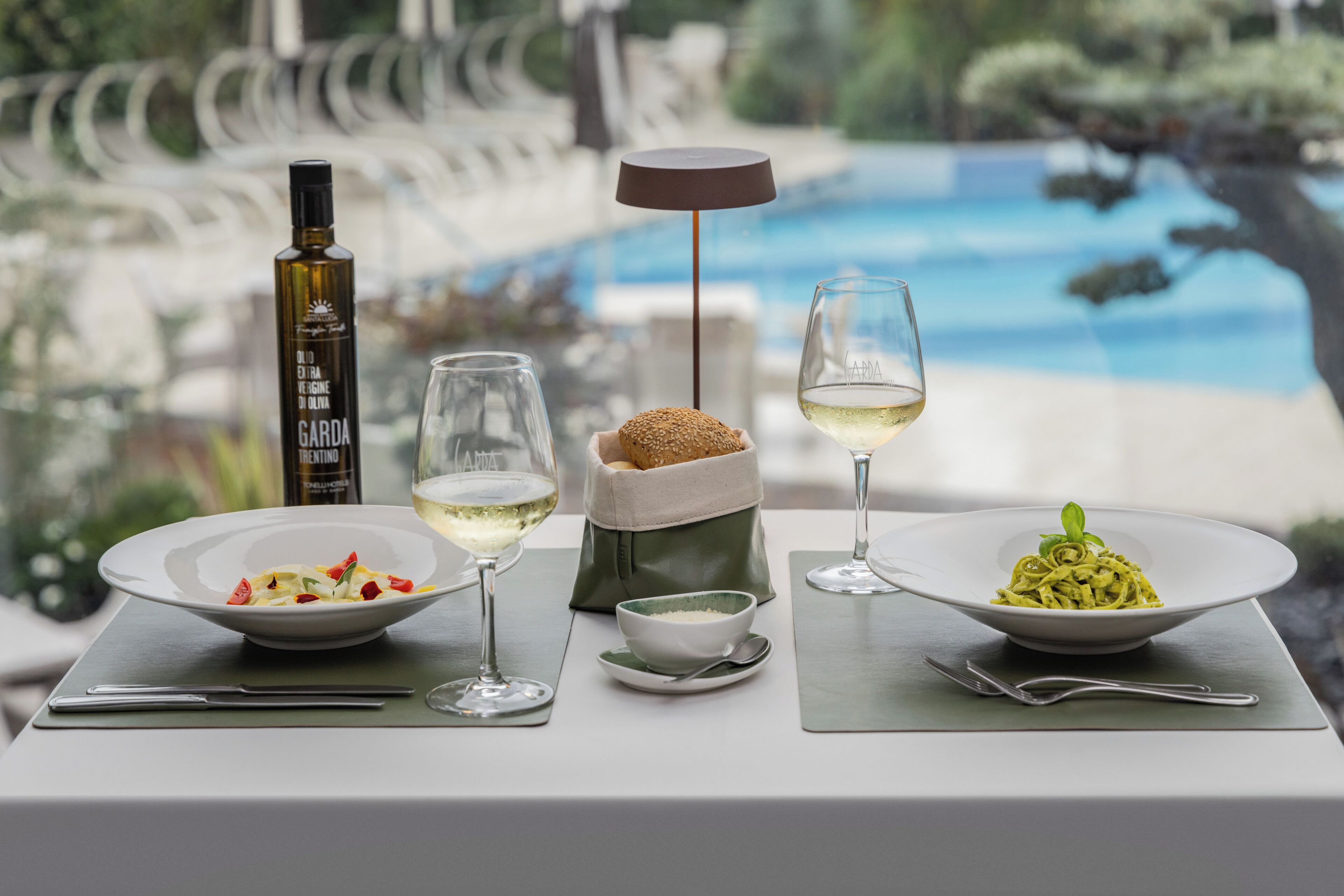 Ein Esstisch mit zwei Pasta-Tellern, einer Olivenölflasche und zwei Gläsern Weißwein, mit Blick auf einen Pool und Grünflächen.