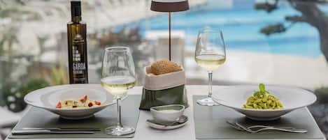 Ein Esstisch mit zwei Pasta-Tellern, einer Olivenölflasche und zwei Gläsern Weißwein, mit Blick auf einen Pool und Grünflächen.