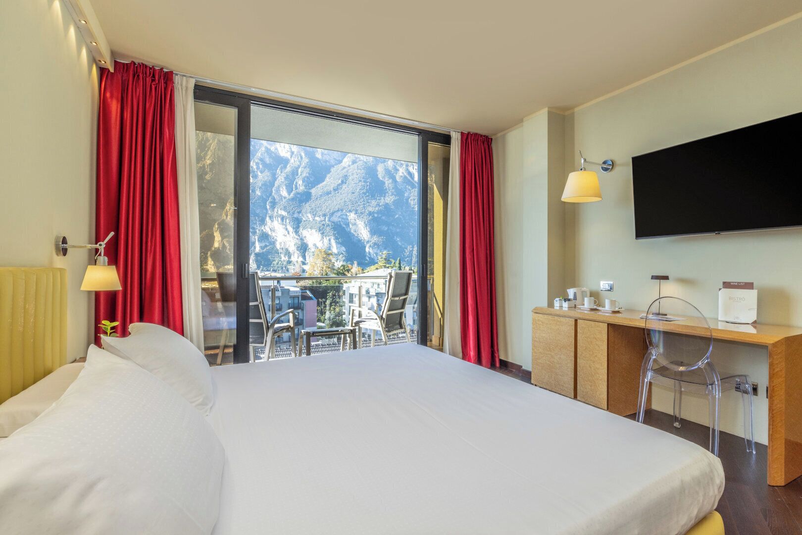 Une chambre d’hôtel avec un grand lit, un bureau avec un ordinateur, une télévision et un balcon offrant une vue sur la montagne.