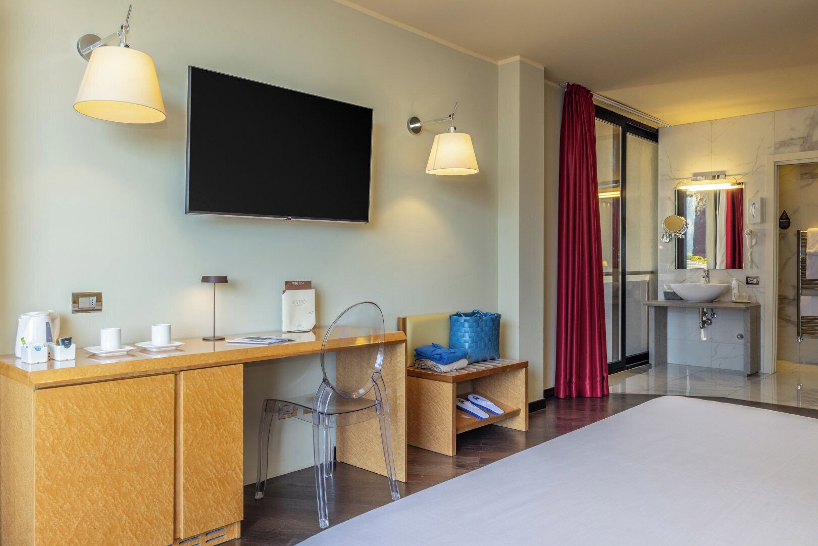 A hotel room with a bed, a desk with a coffee maker, a TV, and a bathroom with a sink and mirror.