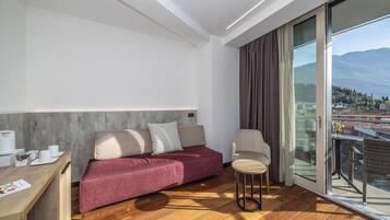 A modern hotel room with a wooden floor, a red sofa, a small round table, a chair, and a large window offering a city view.