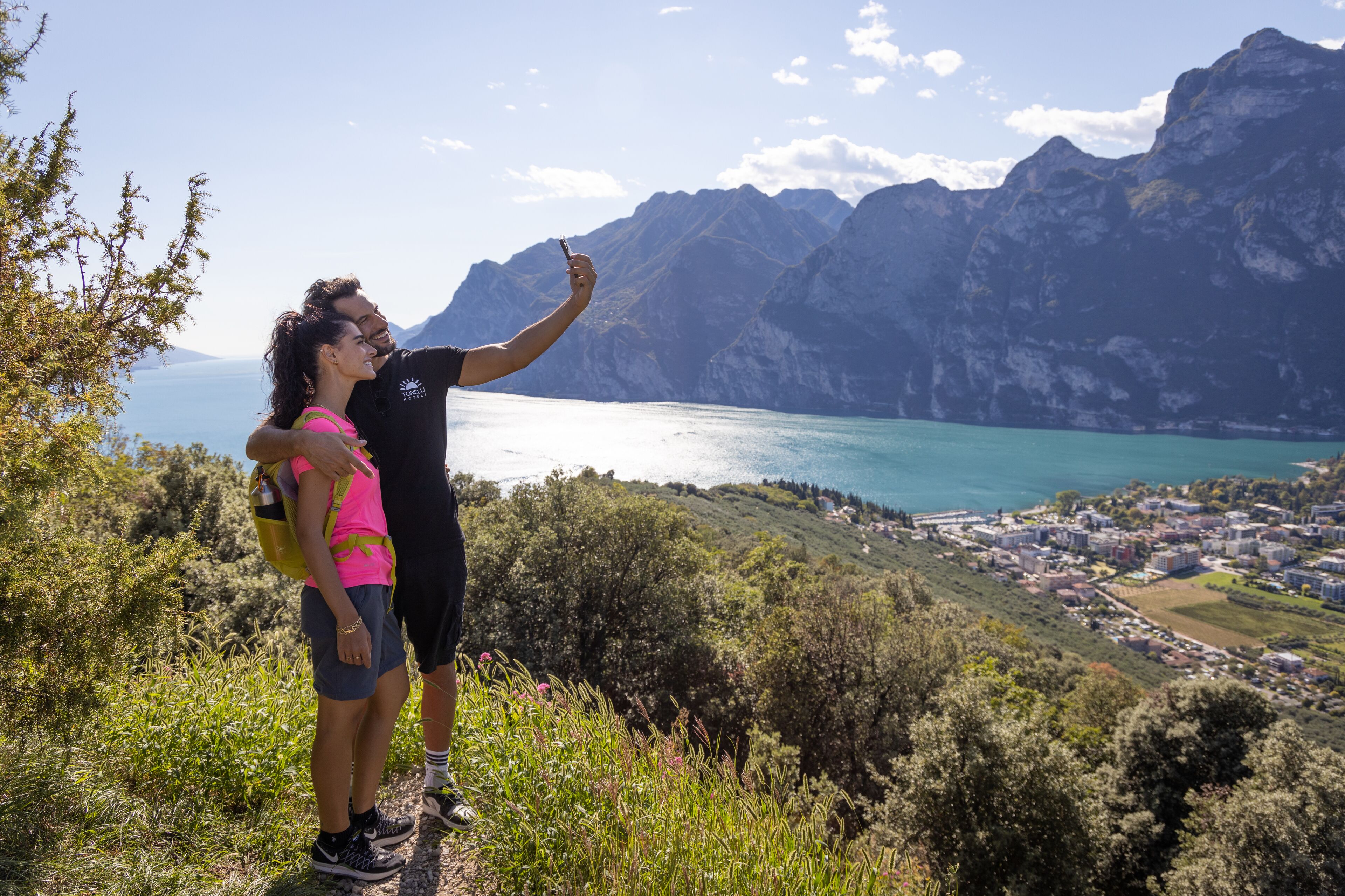 A couple taking a selfie with a mountainous backdrop and a coastal town below.