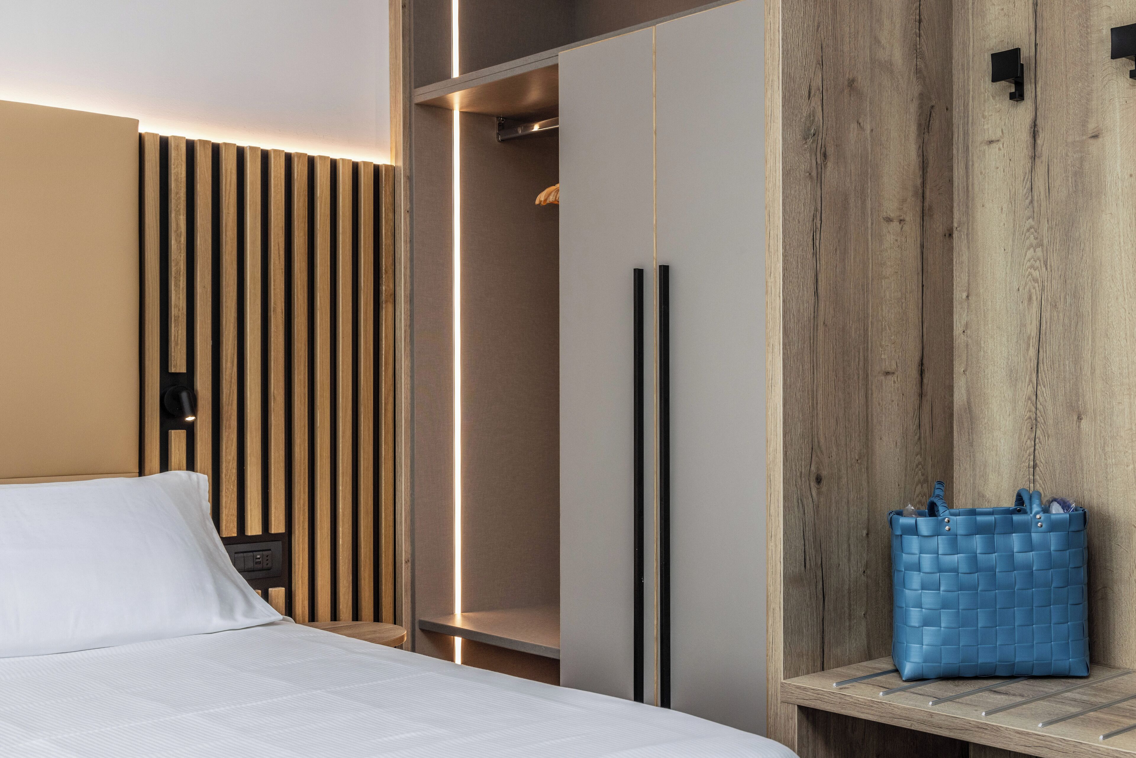 A modern hotel room with a bed, a wardrobe, and a wooden bench with a blue bag.