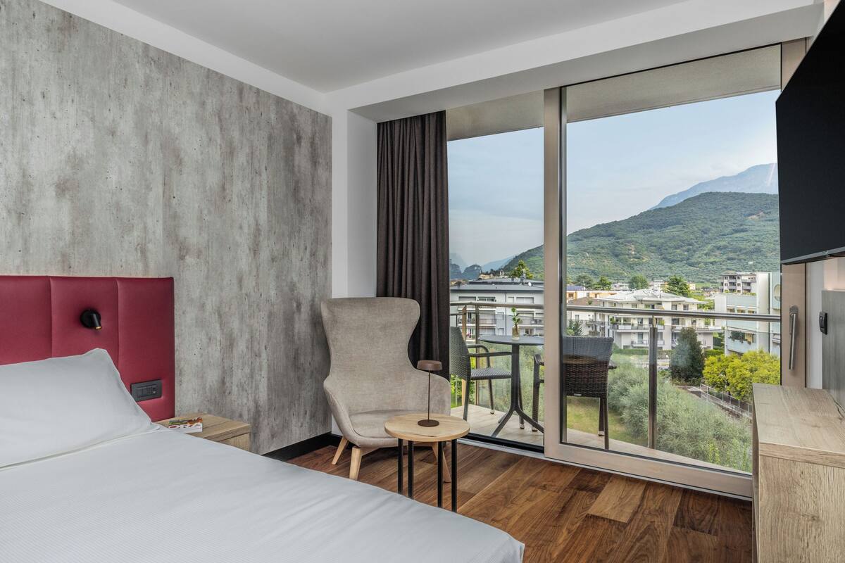 Une chambre d’hôtel moderne avec un grand lit, une tête de lit rouge, un fauteuil gris, une petite table ronde et une vue sur un paysage urbain et des montagnes, le tout visible à travers de grandes fenêtres.