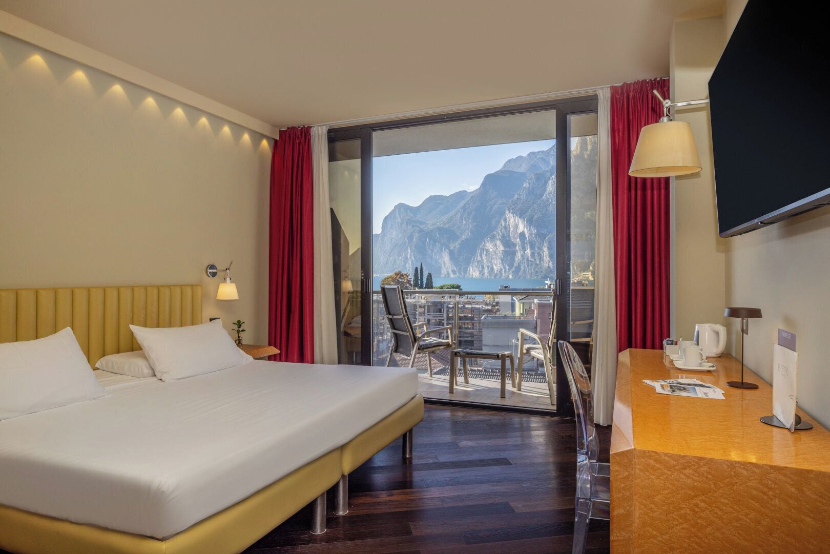 A hotel room with a bed, a desk, a chair, a TV, and a balcony with a mountain view.