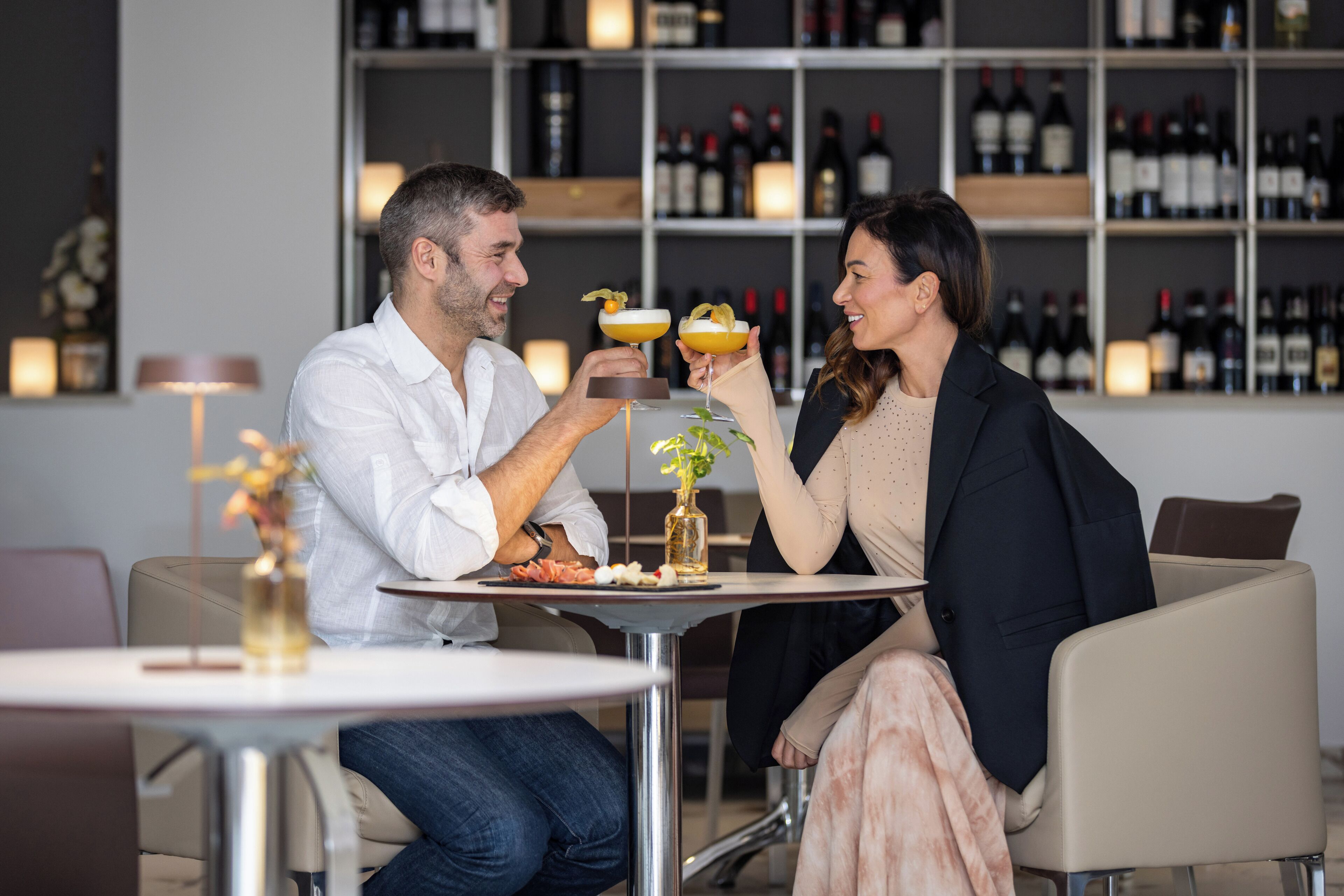 Una pareja sentada en una mesa en un bar moderno, con botellas de vino al fondo.