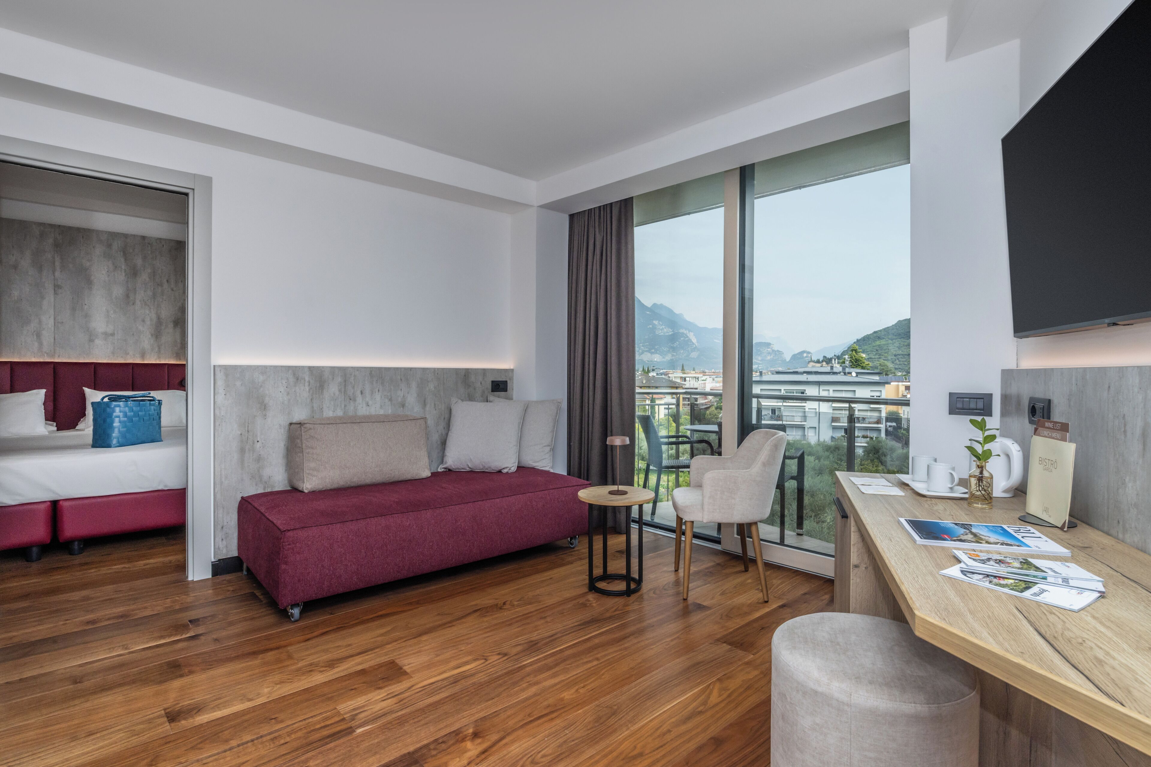 A modern hotel room with a wooden floor, a red sofa, a dining table, and a large window offering a city view.