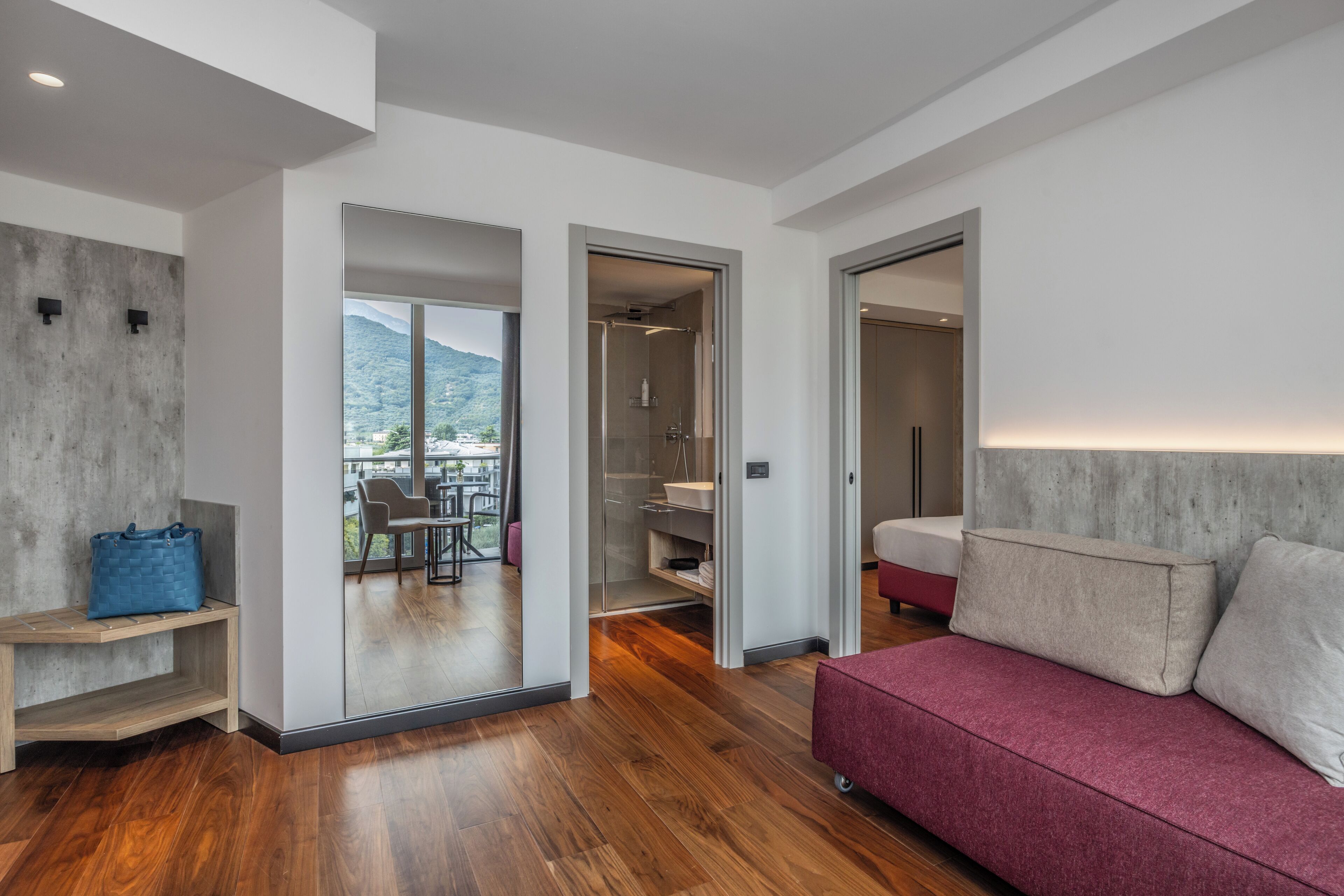 A modern room with a wooden floor, a red sofa, a grey headboard, and a view of a balcony with outdoor furniture.