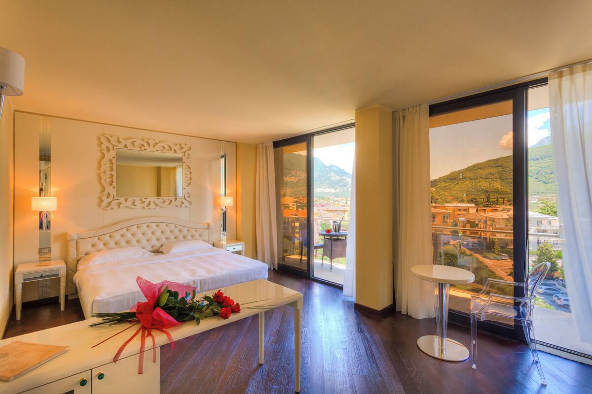 Une chambre d’hôtel avec un grand lit, une table de chevet agrémentée d’un bouquet de fleurs, une petite table ronde et un balcon offrant une vue sur la ville et les montagnes.