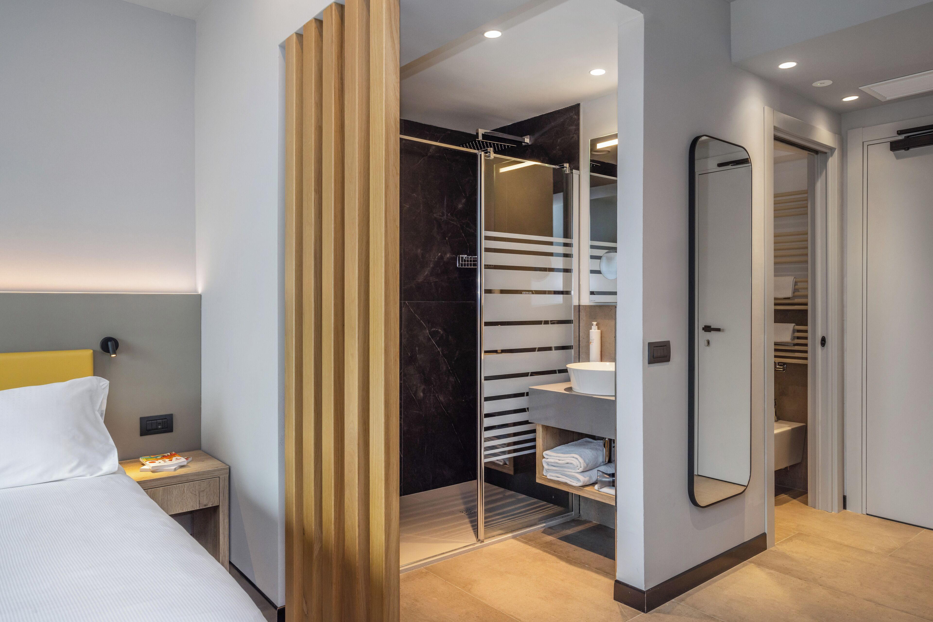 A modern hotel room with a bed, bedside table, and a walk-in closet with a mirror and shelves.