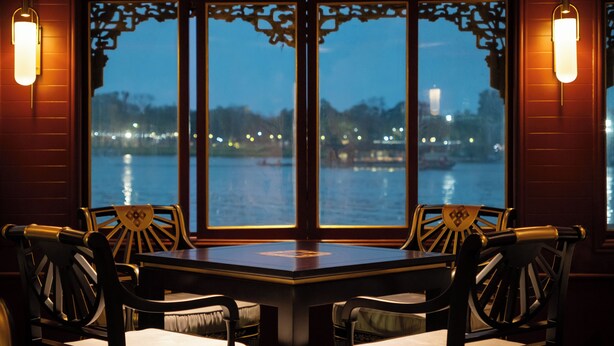 A dining area with a table and chairs, large windows with decorative frames, and wall-mounted lights.