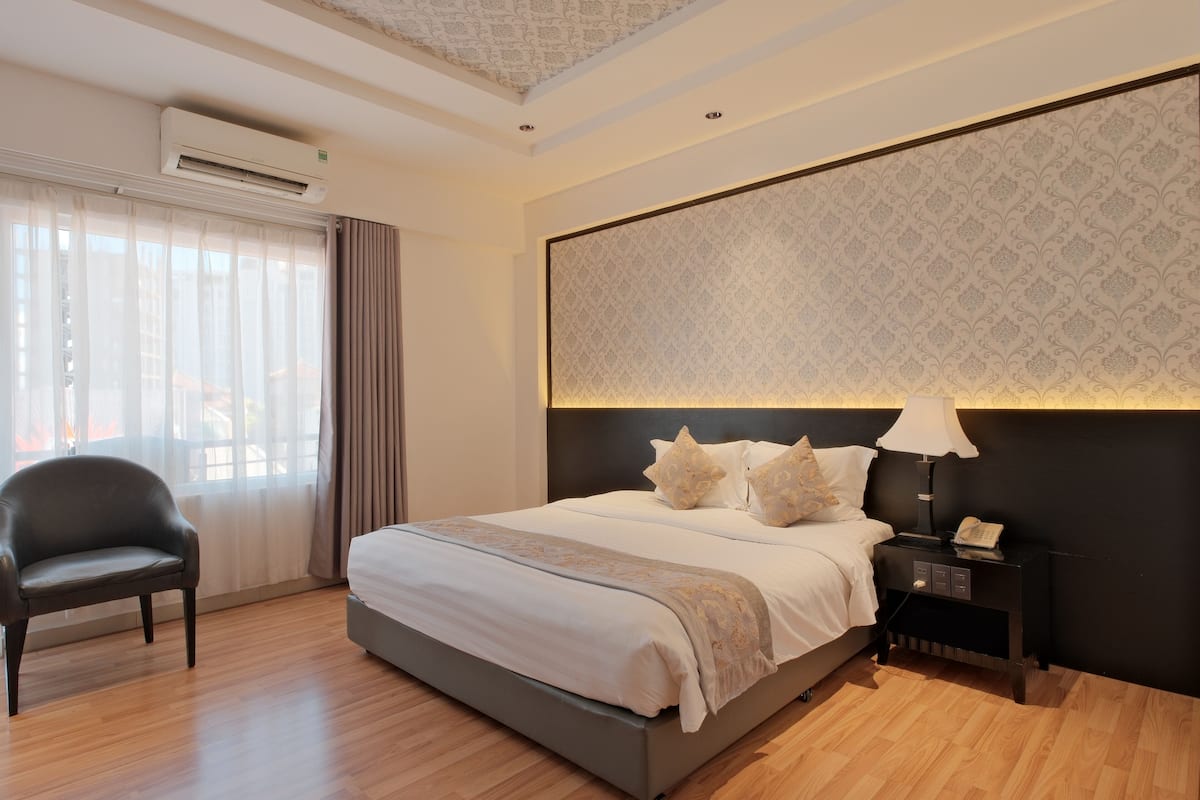 A modern hotel room with a large bed, a bedside table with a lamp, a chair, and an air conditioning unit.