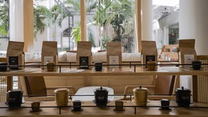 A café with a long table set for a meeting, surrounded by chairs, with a view of palm trees through large windows.