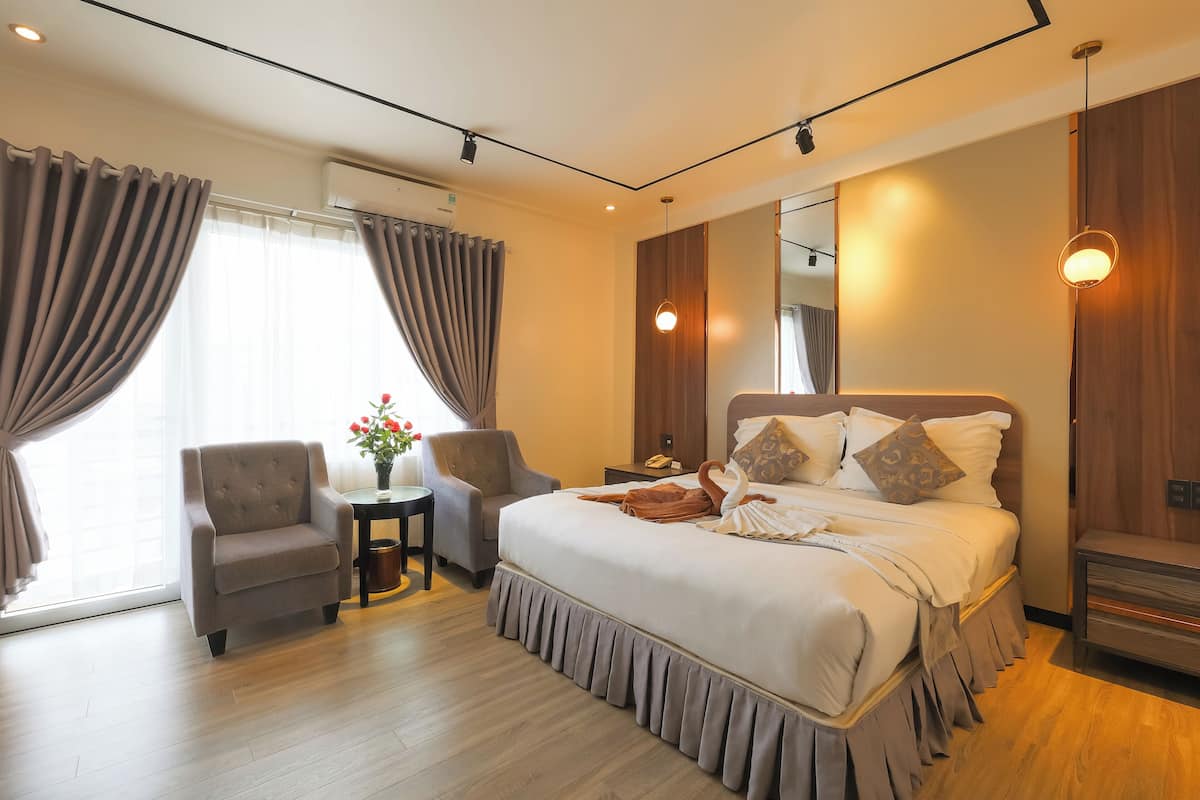 A modern hotel room with a large bed, a sitting area with a chair and table, and a large window with curtains.