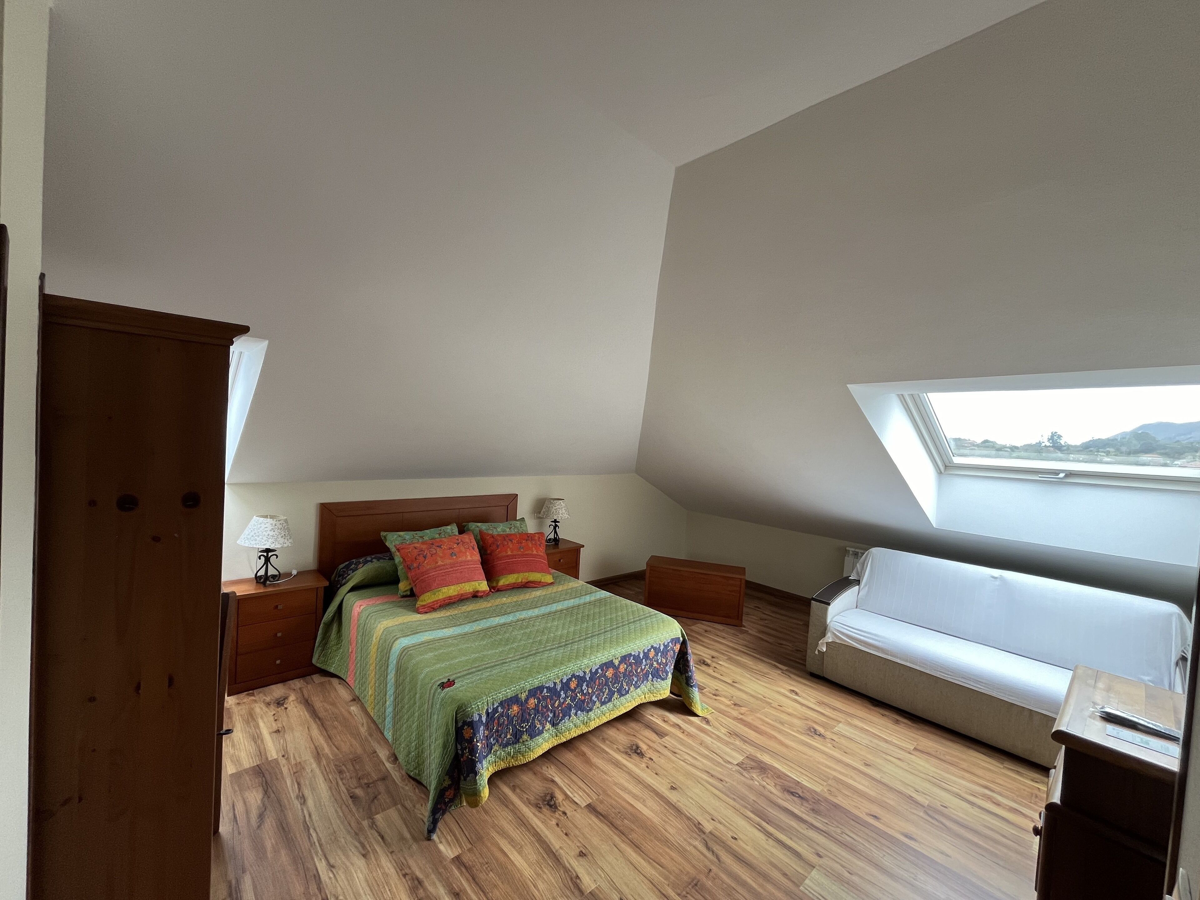 Un dormitorio con techo inclinado, una cama con colcha verde, dos mesitas de noche con lámparas, un armario de madera y un tragaluz.