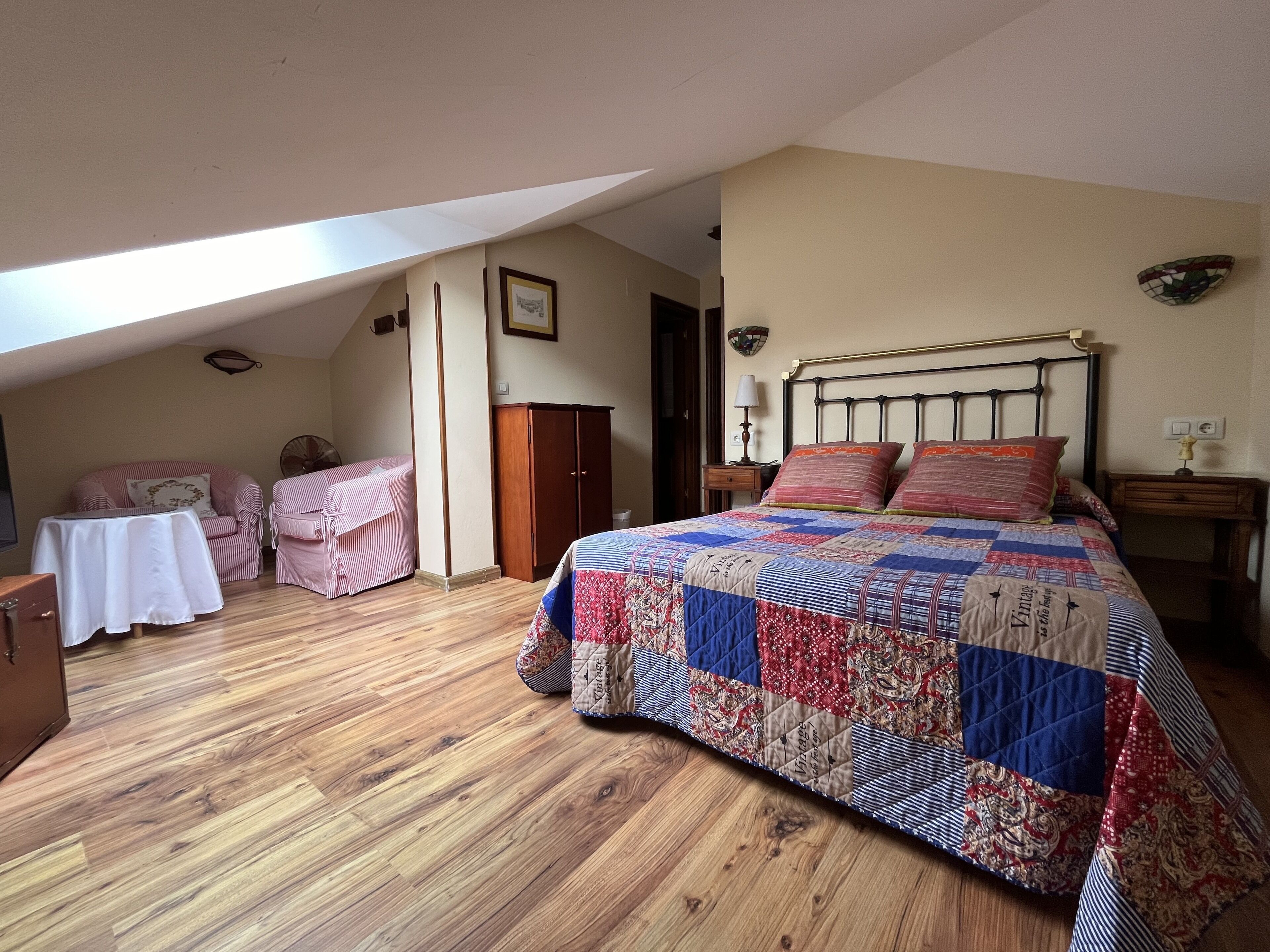 Un dormitorio con una cama grande, suelo de madera, una silla, una mesita y un armario.