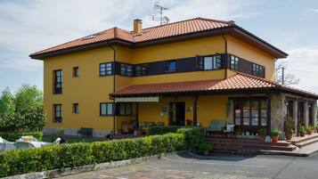 A two-story yellow building with a red-tiled roof, a covered porch with outdoor seating, and a landscaped garden.