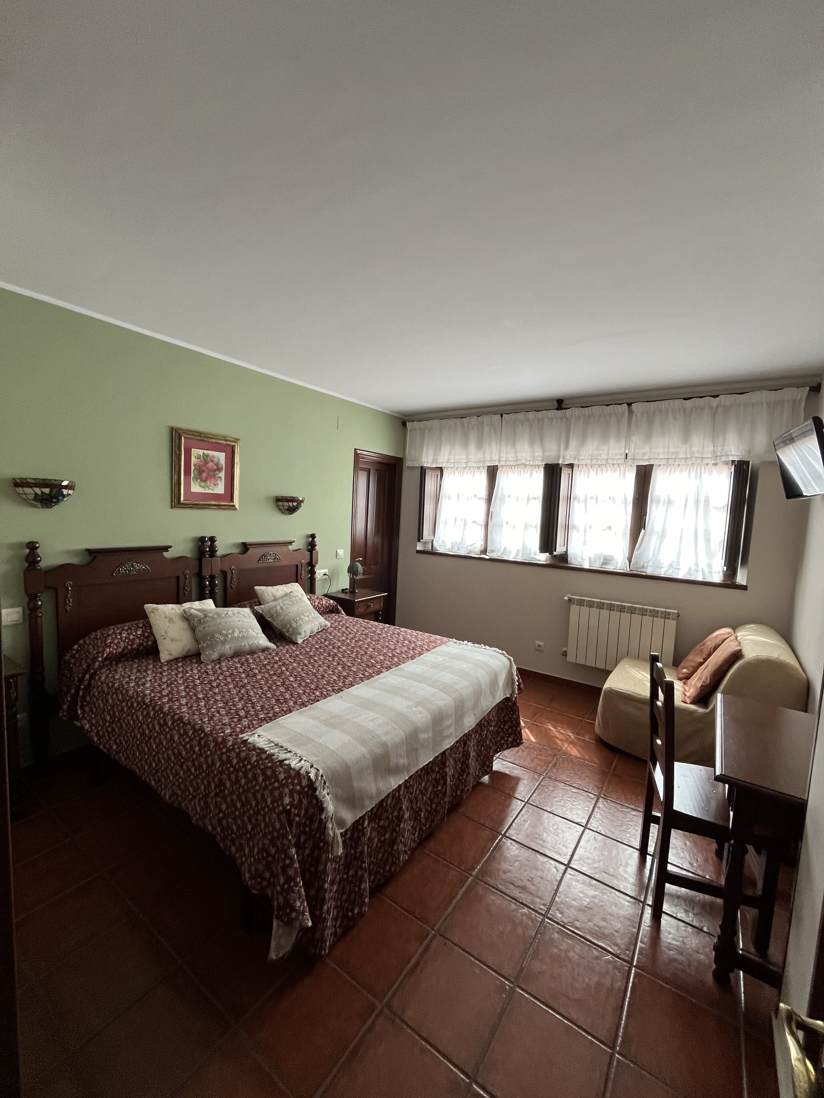 Een slaapkamer met een bed, een stoel, een raam met gordijnen, een radiator en een wandlamp.