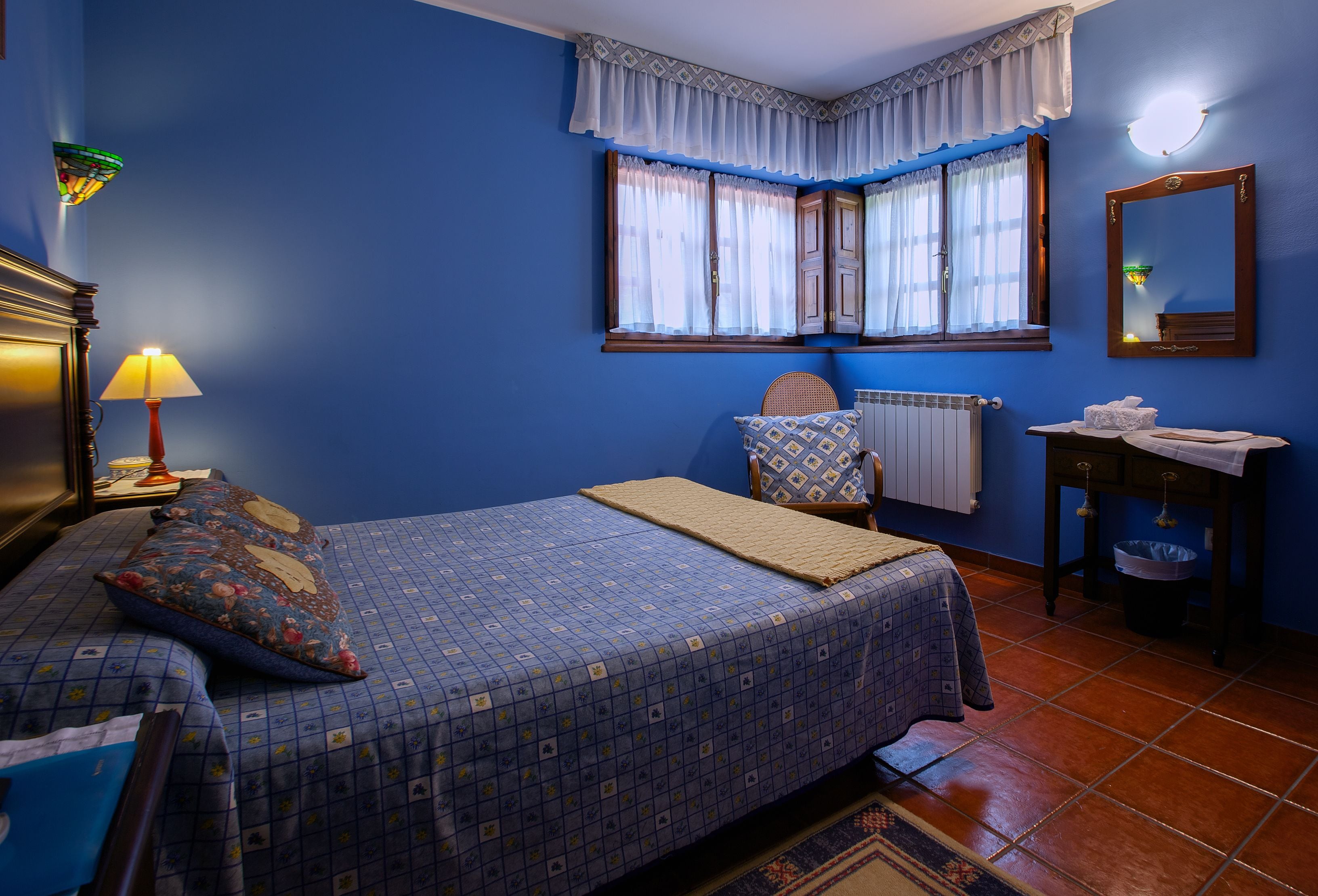 Une chambre à coucher comprenant un lit, une table de chevet, une lampe, une fenêtre avec des rideaux, un radiateur, un miroir et une petite table sur laquelle est posée une serviette.