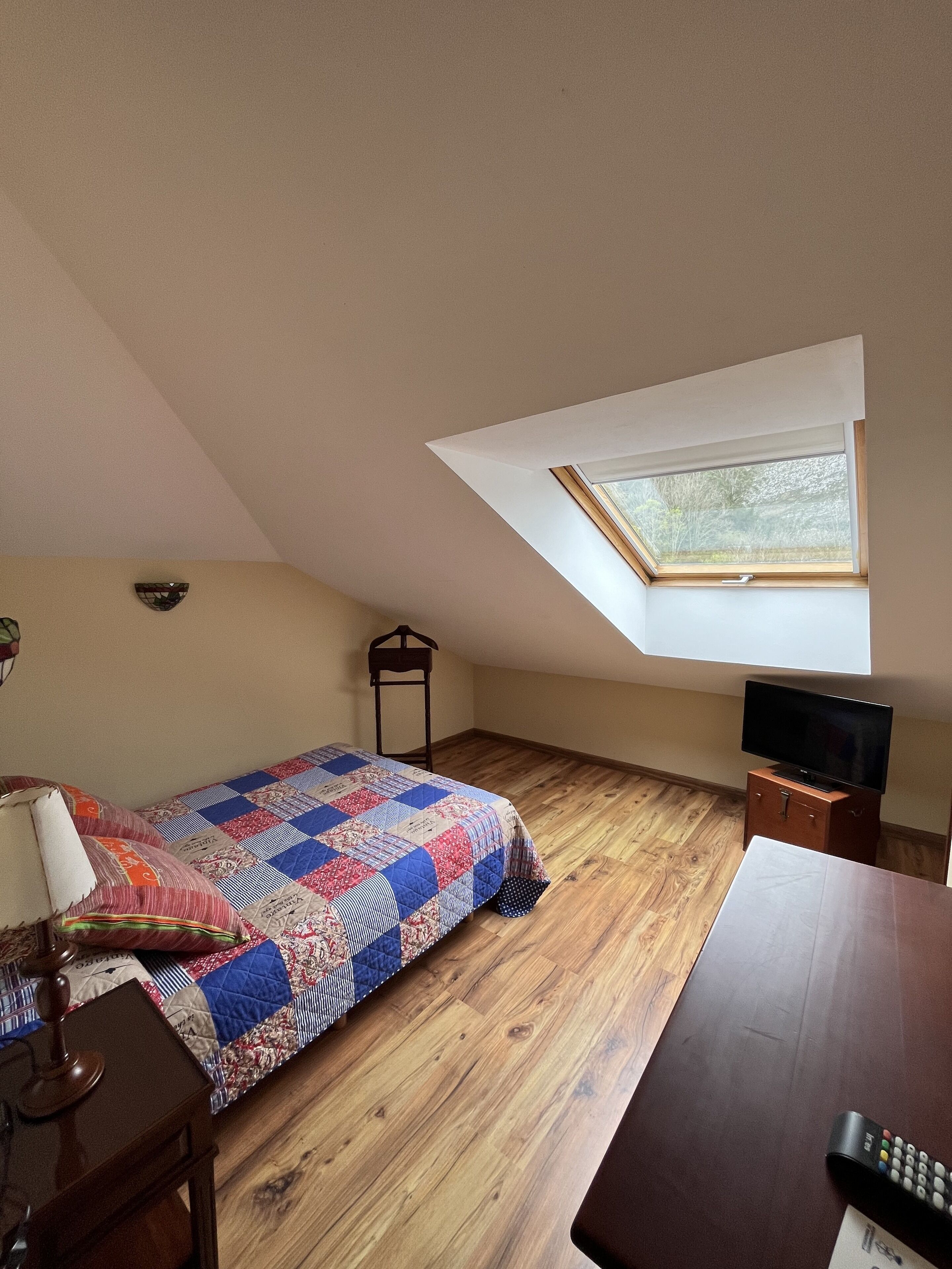 Un dormitorio con techo inclinado, tragaluz, cama con colcha colorida, cómoda de madera, televisor sobre un soporte y una lámpara.