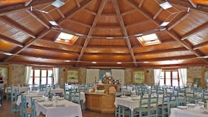 Een ruime eetzaal met meerdere tafels en stoelen, een centraal gelegen bar en een hoog houten plafond met dakramen.